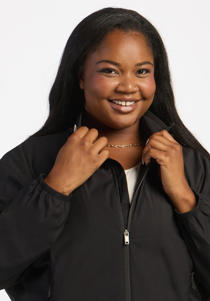 Woman smiling in Woolx Cooper Windbreaker - Black, with zip pockets and gold chain, holding the collar in both hands.