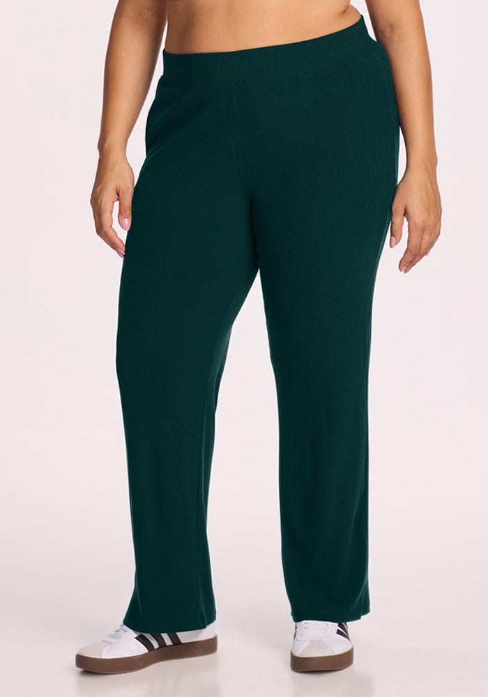 Model wears Woolx Luca Ribbed Pants in Deep Pine with white sneakers, shown from shoulders down on a light background. 