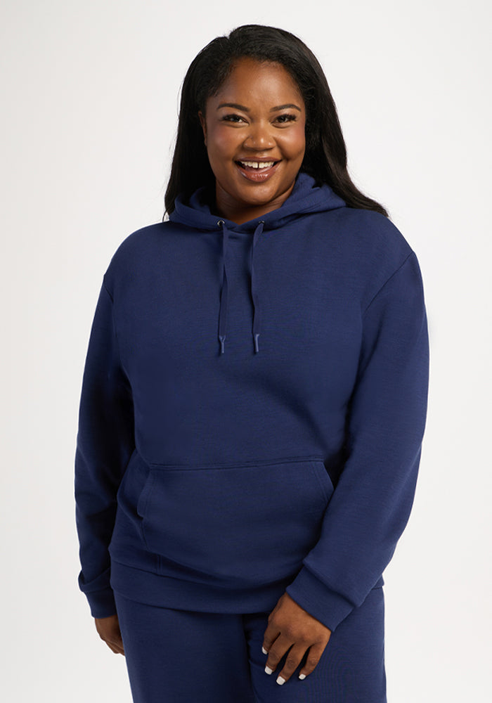Smiling woman in Woolx Avery Hoodie - Ocean Cavern and matching pants, standing against a plain white background. 
