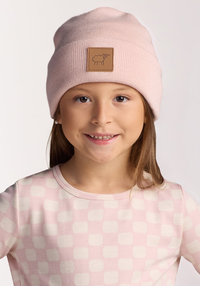 A young girl smiles at the camera, wearing the Woolx Kids Baylor Beanie - Little Ballerina, a light pink winter hat with a brown patch, and a light pink checkered shirt against a plain white background.