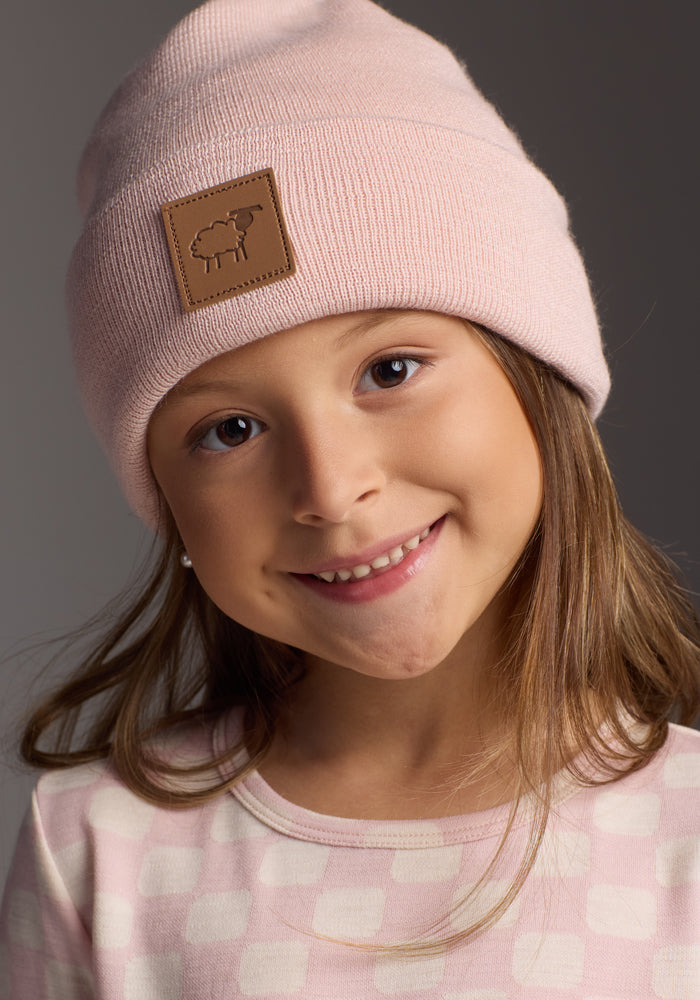 A young girl with long brown hair smiles softly at the camera, wearing the Woolx Kids Baylor Beanie in Little Ballerina and a pink patterned shirt against a neutral background.