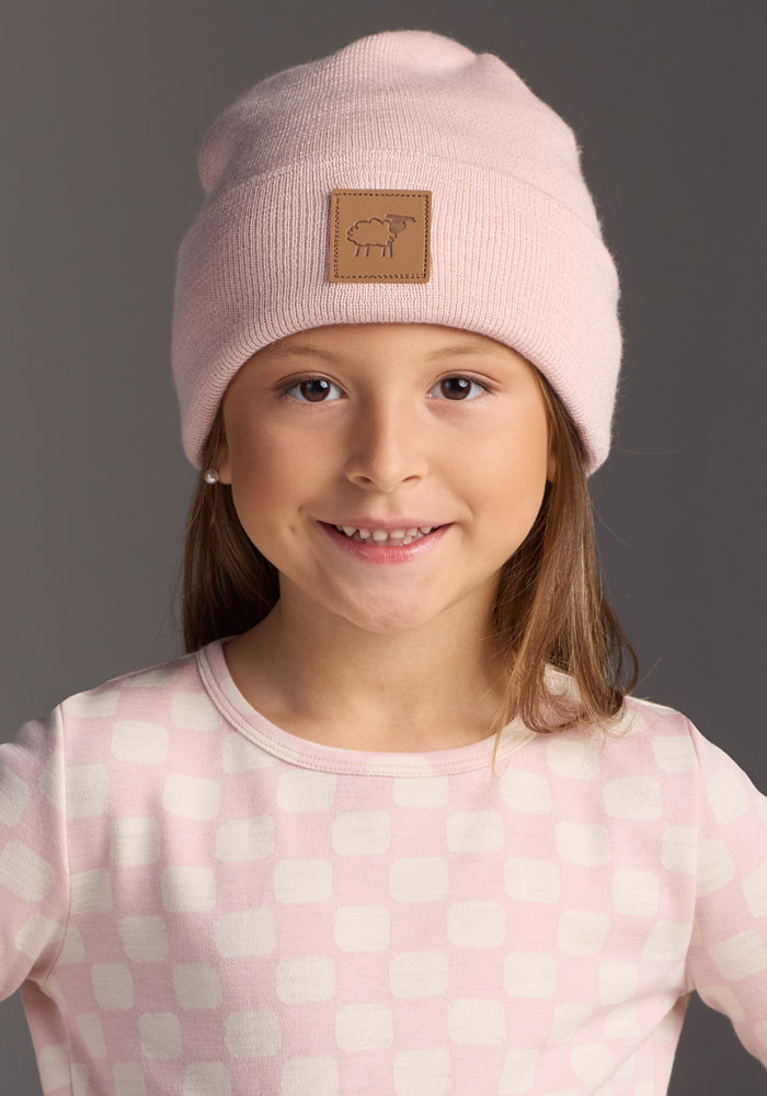 A young girl in a Woolx Kids Baylor Beanie - Little Ballerina and a pink and white checkered shirt smiles at the camera against a plain gray background.