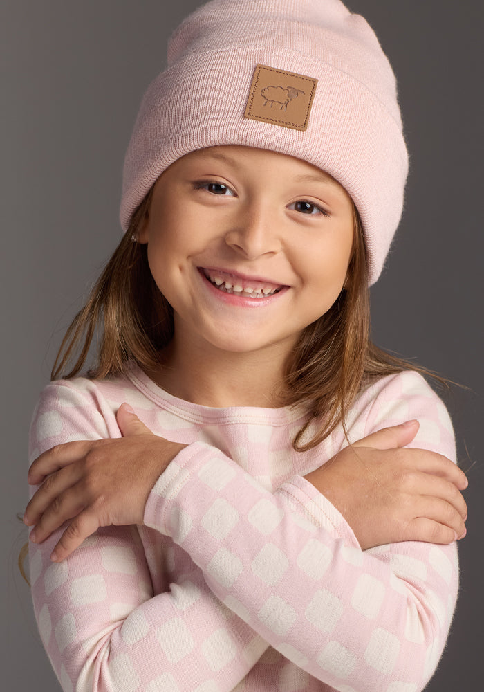 Smiling young girl with long hair, wearing the Woolx Kids Baylor Beanie - Little Ballerina and a light pink checkered long-sleeve shirt, stands with arms crossed against a gray background.