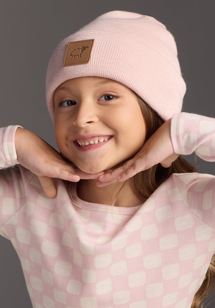 A young child smiles with hands under their chin, wearing the Woolx Kids Baylor Beanie in Little Ballerina and a matching pink geometric long-sleeve shirt, set against a gray background.