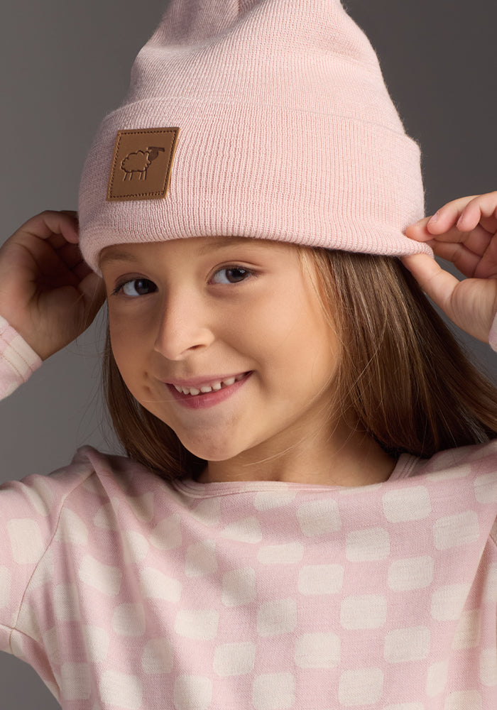 A young girl smiles while wearing the Woolx Kids Baylor Beanie - Little Ballerina, a light pink stretchy hat with a sheep patch, paired with a matching pink patterned long-sleeve shirt. She holds her beanie with both hands against a neutral background.