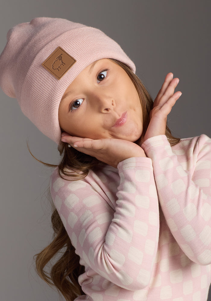 A young girl poses playfully with her hands framing her face, wearing the Woolx Kids Baylor Beanie - Little Ballerina and a pink patterned long-sleeve shirt against a plain gray background.