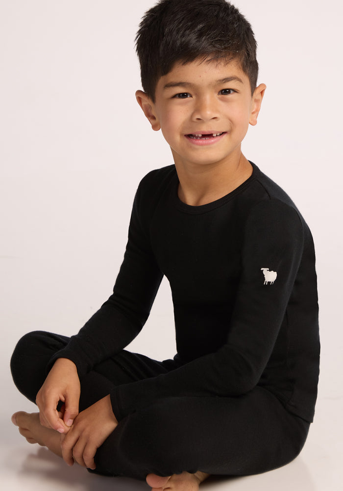 A young child with short dark hair sits cross-legged, smiling at the camera while wearing the Woolx Kids Explorer Base Layer Top in black, featuring a small white sheep logo on the sleeve, set against a plain light background.