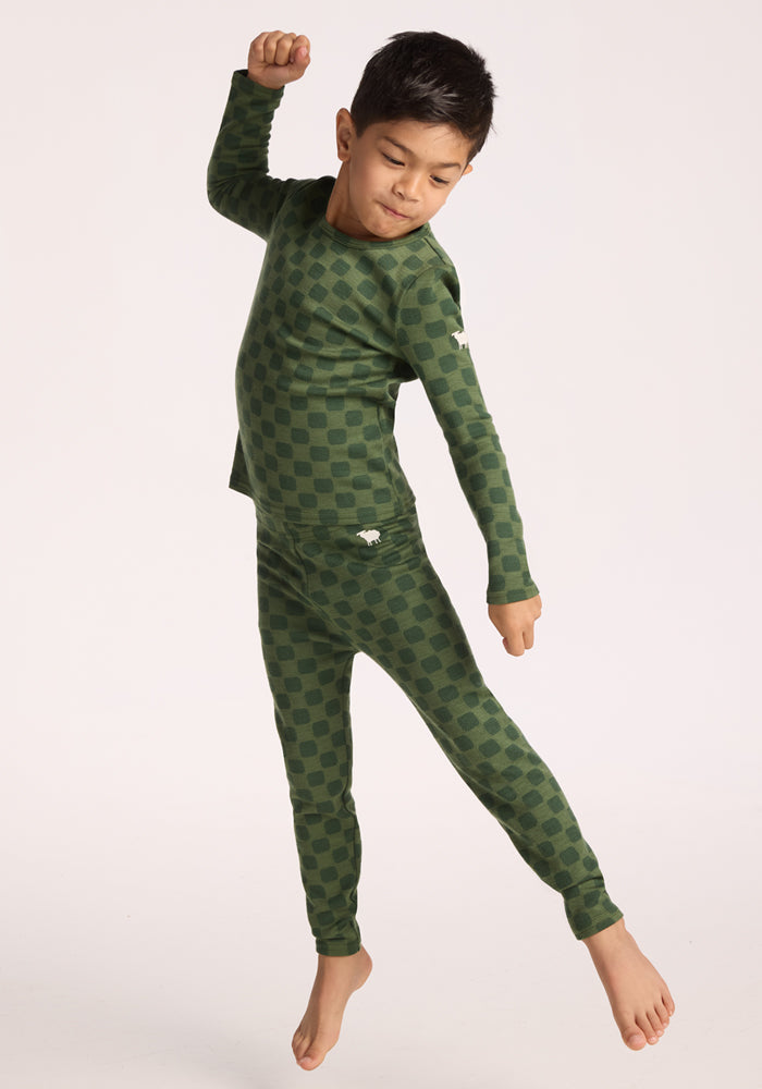 A young child wearing Woolx Kids Explorer Base Layer Bottoms - Frog Hop jumps joyfully in the air against a plain, light background.