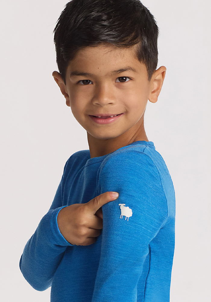 A young boy with dark hair smiles and points to a small white sheep logo on the sleeve of his bright blue Woolx Kids Explorer Base Layer Top in Blue Razz, against a plain light background.