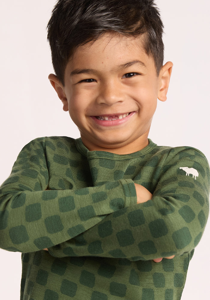 Smiling young boy with dark hair wears the Woolx Kids Explorer Base Layer Top - Frog Hop, a green long-sleeve Merino wool shirt with a checkered pattern and white sheep logo on the sleeve. Comfortable, breathable, perfect for kids.