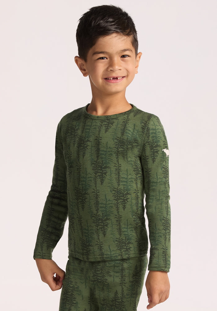 A young boy with short dark hair smiles at the camera, wearing the Woolx Kids Explorer Base Layer Top - Forest Friends and matching pants. The background is plain and light-colored. 