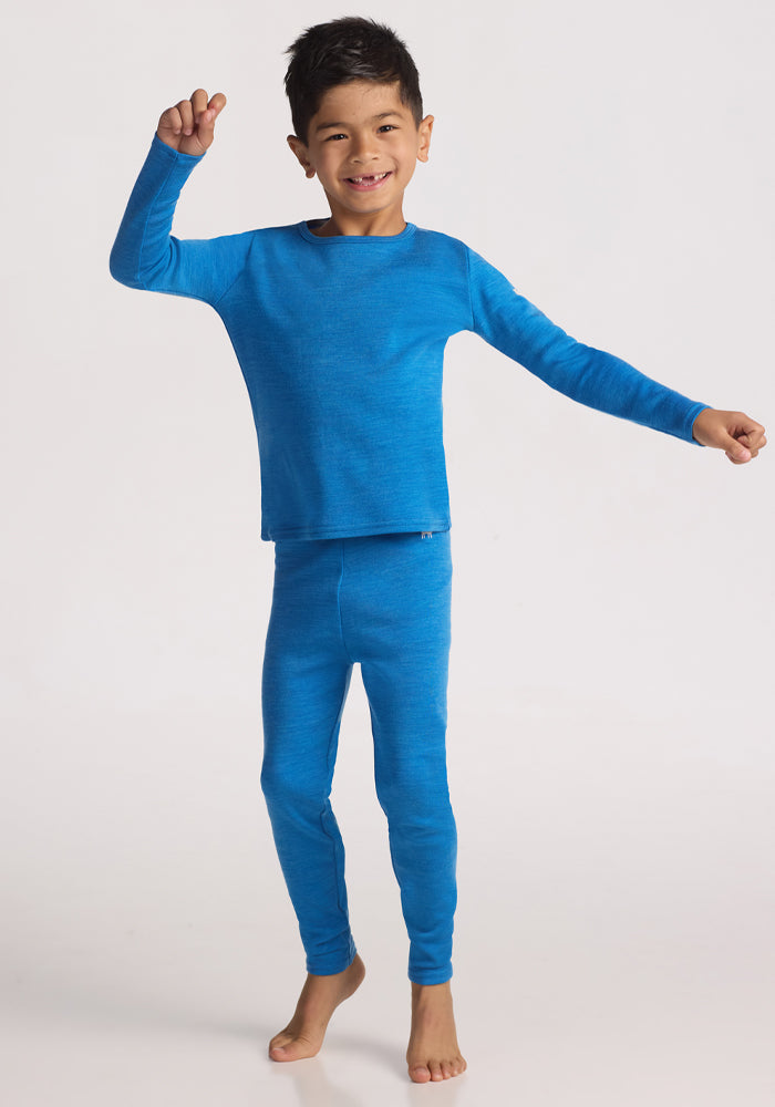 A smiling young boy stands barefoot on a white background, showing off his cozy Woolx Kids Explorer Base Layer Bottoms in Blue Razz, paired with a matching merino wool thermal top, one arm raised and the other out to the side.