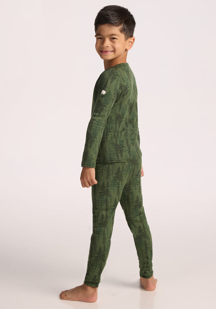 A young boy smiles over his shoulder, barefoot in matching green Woolx Kids Explorer Base Layer Bottoms - Forest Friends, featuring a dark green tree pattern—perfect cozy merino wool pajamas for chilly nights against a plain backdrop.