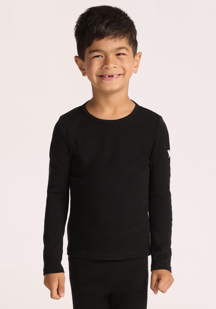 A young boy with dark hair smiles against a plain light background, wearing the Woolx Kids Explorer Base Layer Top in black, crafted from moisture-wicking Australian Merino wool, paired with matching black pants. 