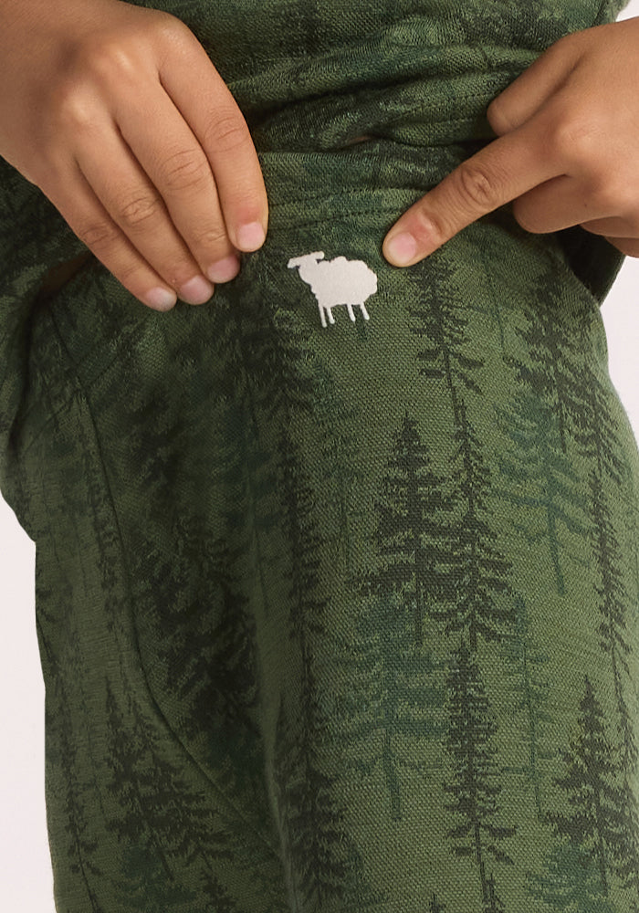 A person points to a white sheep graphic on Woolx Kids Explorer Base Layer Bottoms - Forest Friends, featuring green merino wool fabric with a dark green tree pattern.