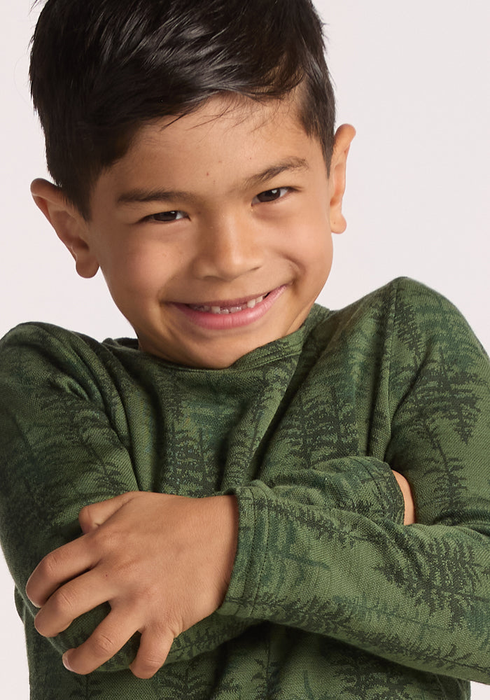 A young boy with short dark hair smiles warmly, arms crossed, wearing the Woolx Kids Explorer Base Layer Top - Forest Friends. This green, long-sleeved merino wool shirt has a subtle tree branch pattern. The background is plain and light-colored.