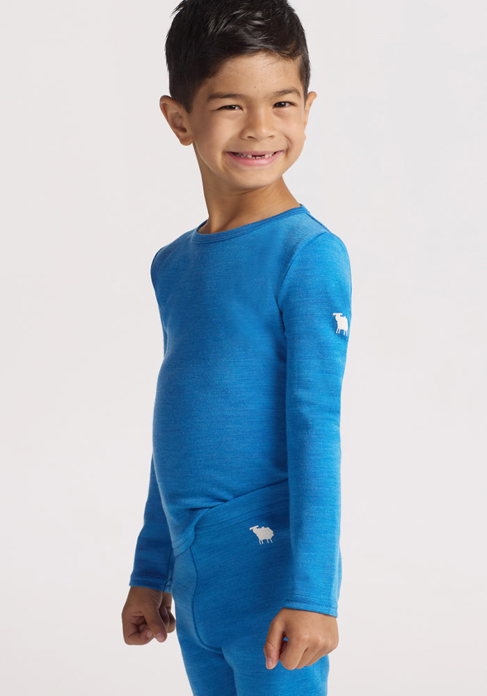 A young child with short dark hair smiles, standing sideways in a Woolx Kids Explorer Base Layer Top - Blue Razz with small white sheep logos on the sleeve, paired with matching pants, against a plain light background.