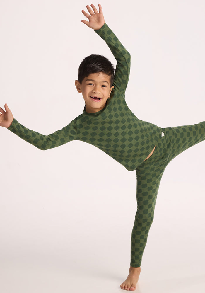 A young child in matching green checkered pajamas and the Woolx Kids Explorer Base Layer Top - Frog Hop balances on one leg with arms outstretched and a joyful expression, posing playfully against a plain light background.