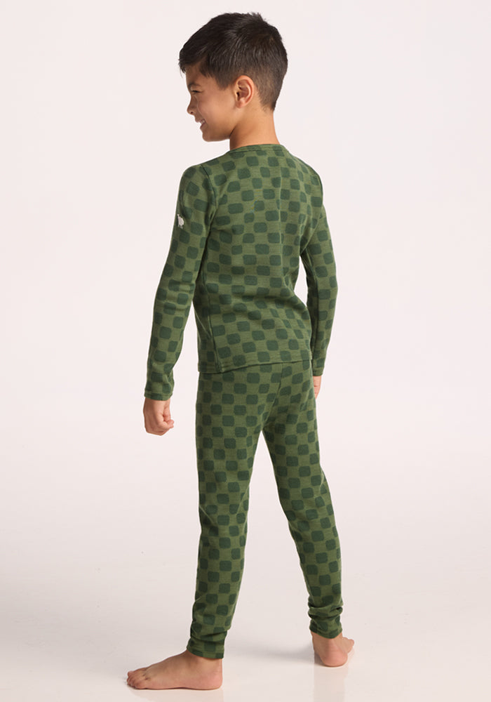 A young boy stands barefoot, facing away, wearing a matching green checkered pajama set with long sleeves and Woolx Kids Explorer Base Layer Bottoms - Frog Hop. The background is plain white.