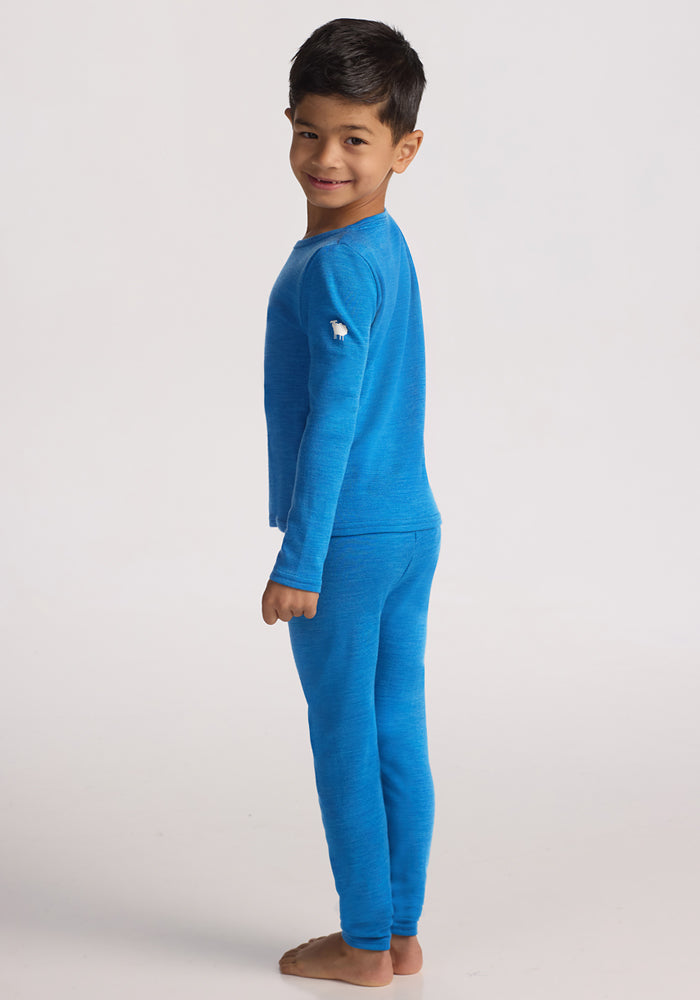 A young boy with dark hair smiles barefoot, facing sideways in Woolx Kids Explorer Base Layer Bottoms - Blue Razz, paired with a matching bright blue merino wool top, against a plain light background.