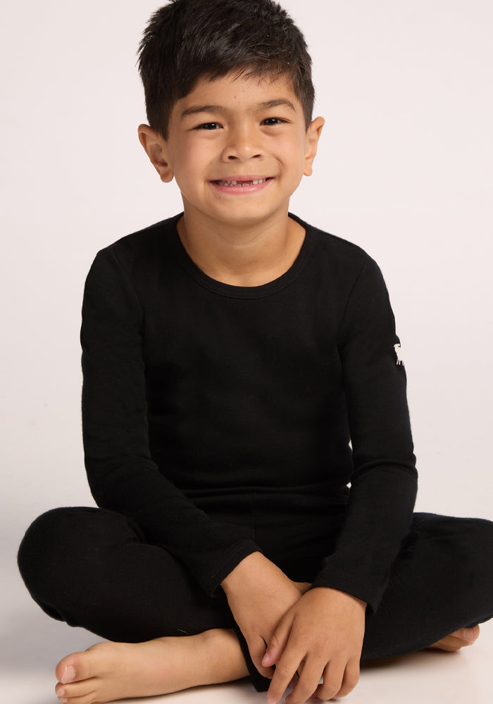 A young boy with short dark hair, smiling at the camera, sits cross-legged on the floor wearing the Woolx Kids Explorer Base Layer Top in black—made from moisture-wicking Australian Merino wool—against a plain light background.