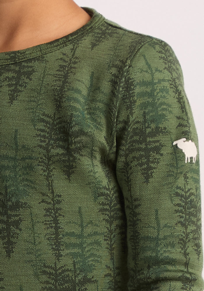 A close-up of the Woolx Kids Explorer Base Layer Top - Forest Friends, a green long-sleeve merino wool top for children featuring a dark green tree pattern and an embroidered white sheep logo on the upper sleeve. Part of a child’s neck and shoulder is visible.