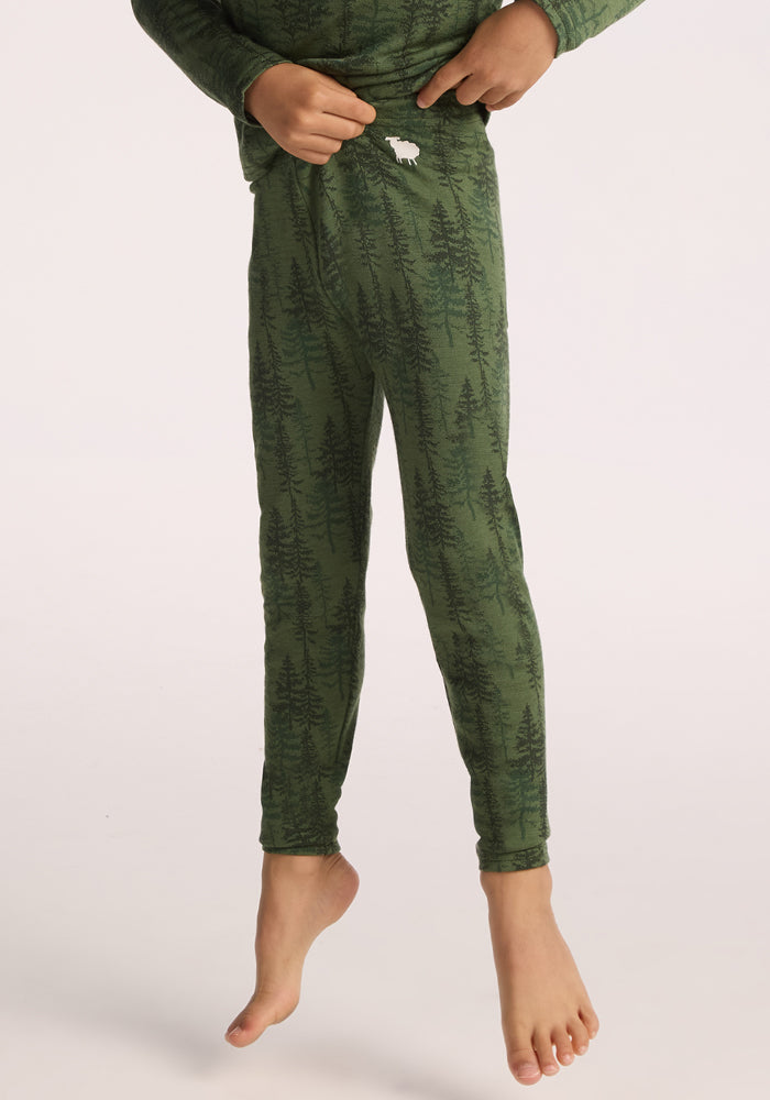 A child wearing Woolx Kids Explorer Base Layer Bottoms - Forest Friends, featuring green pajama pants with a tree pattern and a small white moose on the waistband, stands on one leg against a plain background. 