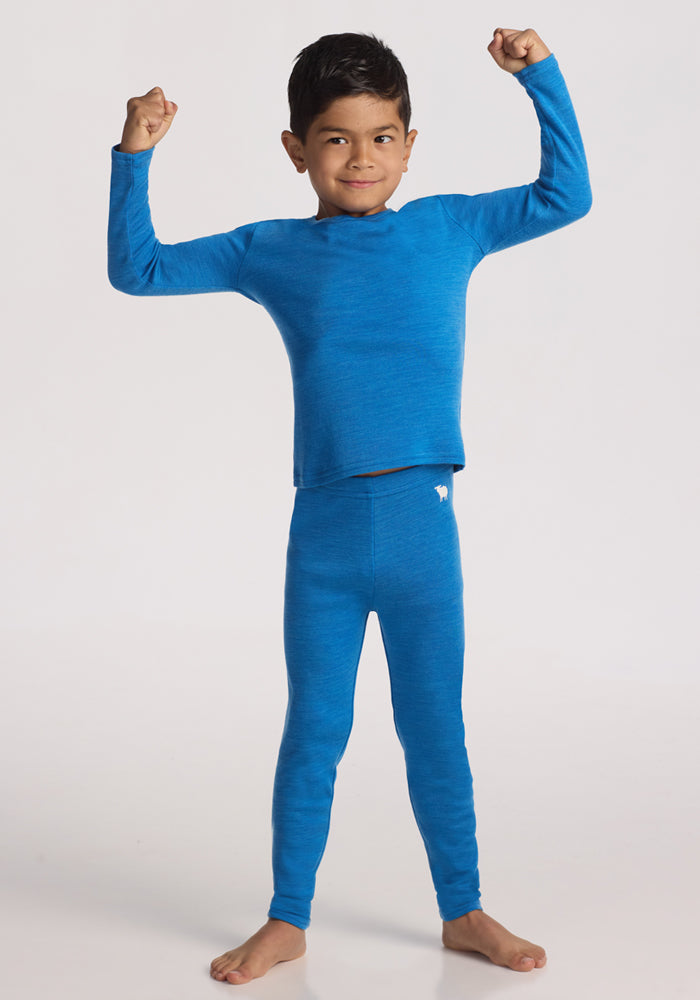 A young boy stands barefoot on a white background, smiling and flexing his arms while wearing Woolx Kids Explorer Base Layer Bottoms in Blue Razz, paired with a matching merino wool top.