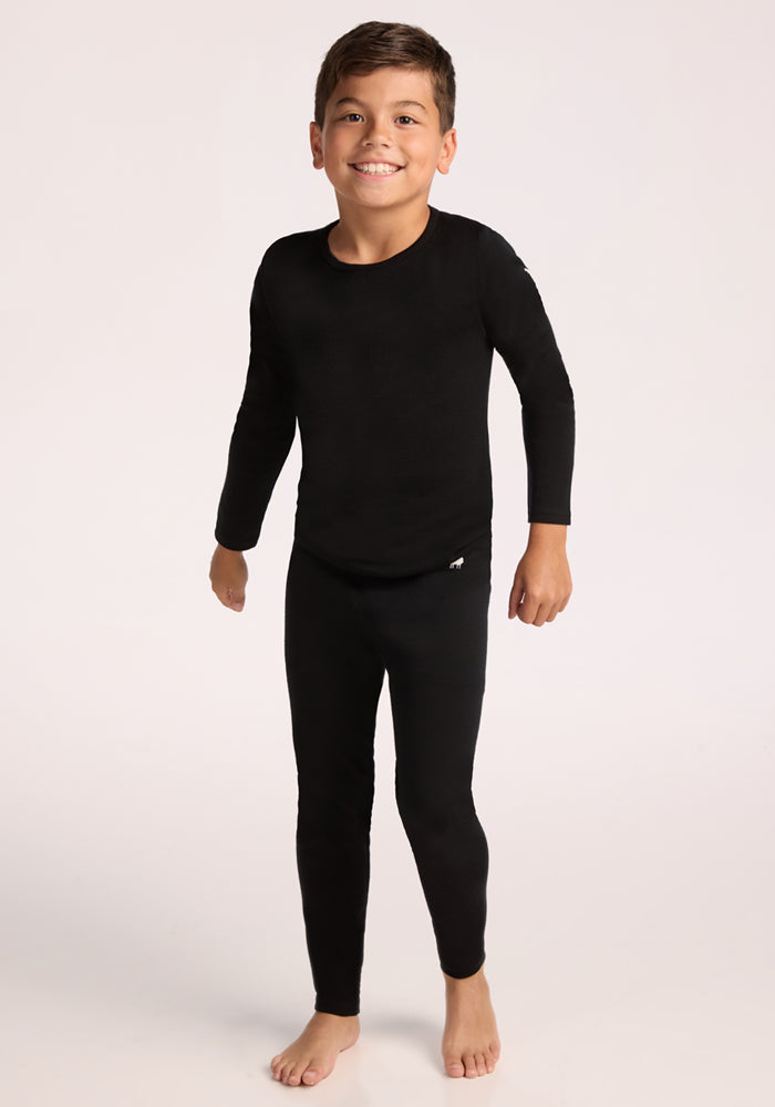 A young boy smiles barefoot against a plain light background, wearing matching black Woolx Kids Explorer Base Layer Bottoms and a long-sleeve top.