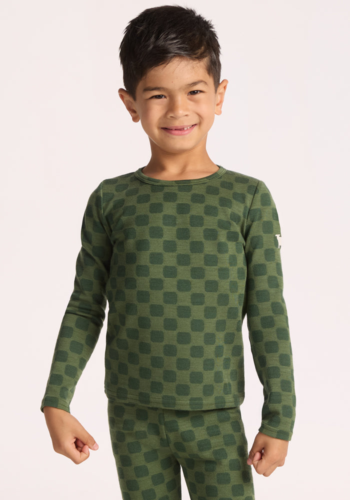 A smiling young child with dark hair wears the Woolx Kids Explorer Base Layer Top - Frog Hop, a cozy and breathable Merino wool thermal featuring a green checkered pattern, standing against a plain light background. 