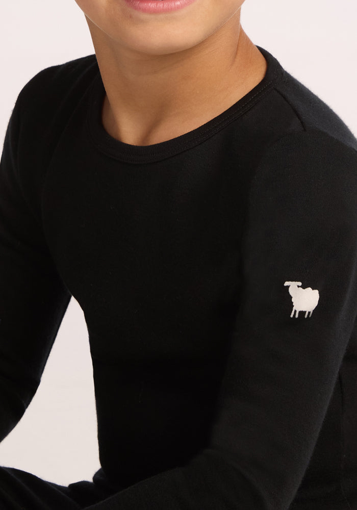 A child in a black Woolx Kids Explorer Base Layer Top made from Australian Merino wool, featuring a small white sheep logo on the left shoulder. The child’s face is partially visible against a plain light background.