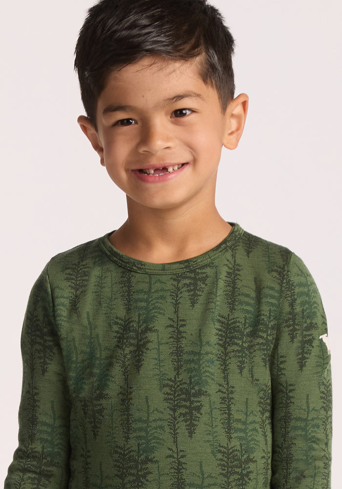 A young boy with short dark hair smiles, showing a gap where he lost a tooth. He wears the Woolx Kids Explorer Base Layer Top - Forest Friends, featuring a leafy green pattern, standing against a plain light background.