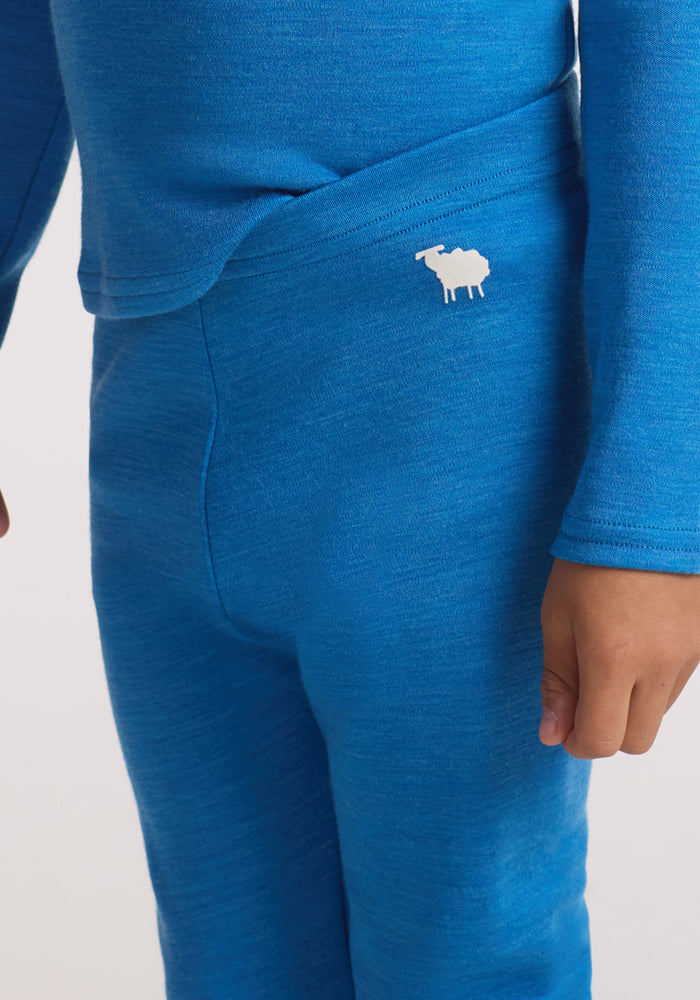 A child in Woolx Kids Explorer Base Layer Bottoms - Blue Razz, showing only their lower torso and arms, with a white moose logo on the waistband, stands against a plain background.
