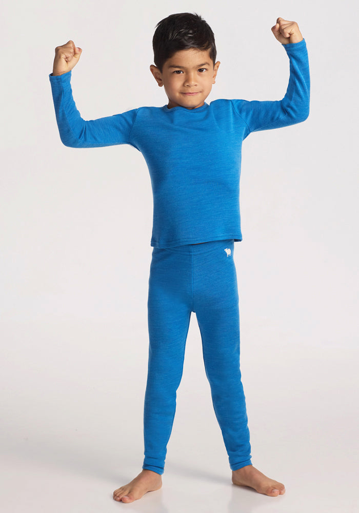 A young child stands barefoot against a plain background, wearing the Woolx Kids Explorer Base Layer Top in Blue Razz and matching leggings, smiling slightly and flexing both arms with fists raised in a strong pose.
