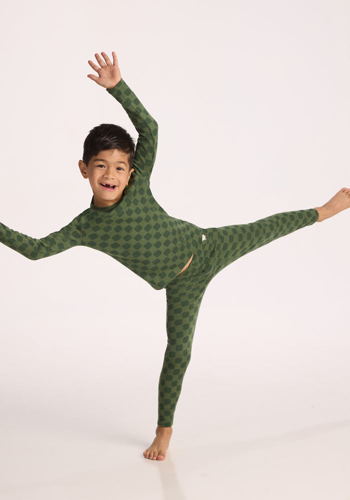 A smiling young boy in green checkered pajamas, wearing Woolx Kids Explorer Base Layer Bottoms - Frog Hop, balances playfully on one leg with arms outstretched against a plain white background.