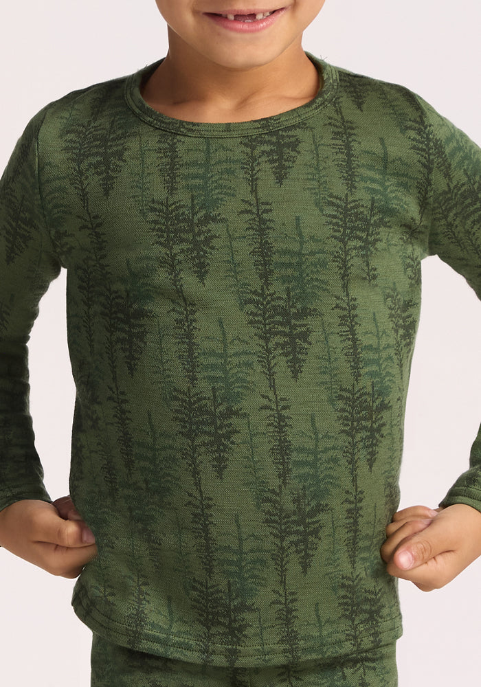 A smiling child wears the Woolx Kids Explorer Base Layer Top - Forest Friends, a long-sleeve green merino wool shirt with a fern leaf design, standing with hands on hips against a plain background.