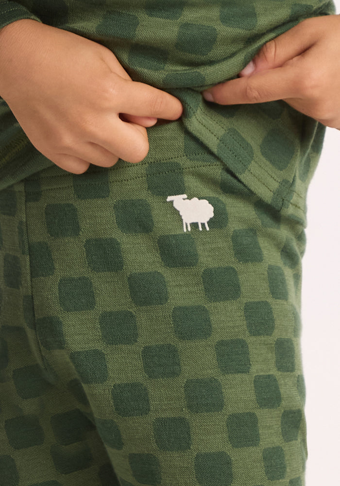 A child's hands adjust the waistband of Woolx Kids Explorer Base Layer Bottoms - Frog Hop, featuring green checkered wool and a small white sheep design near the pocket.