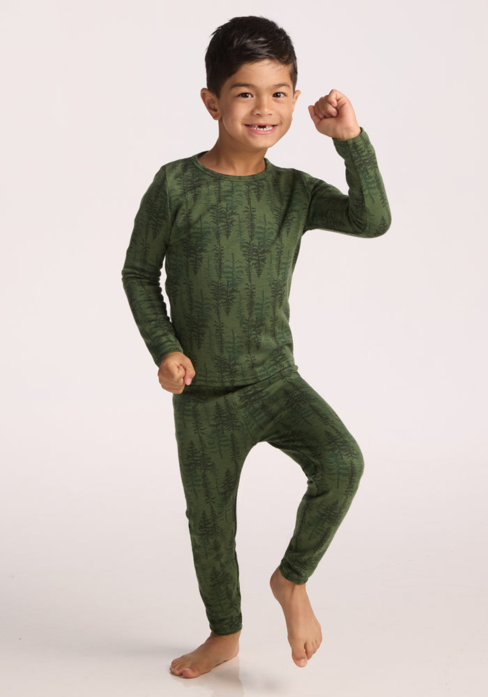 A young boy wearing Woolx Kids Explorer Base Layer Bottoms - Forest Friends stands on one foot, smiling and raising an arm playfully against a plain light background, showing off these comfy kids’ merino wool baselayers.