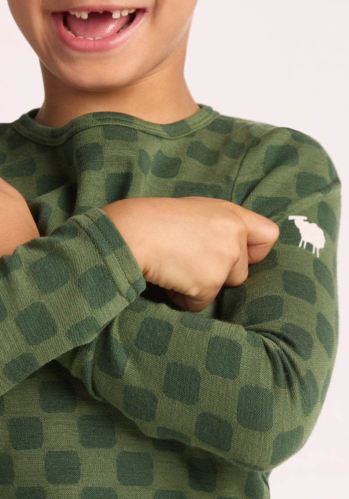 A child smiles with arms crossed, wearing the Woolx Kids Explorer Base Layer Top - Frog Hop, a green checkered wool thermal featuring a small white sheep logo on the sleeve.