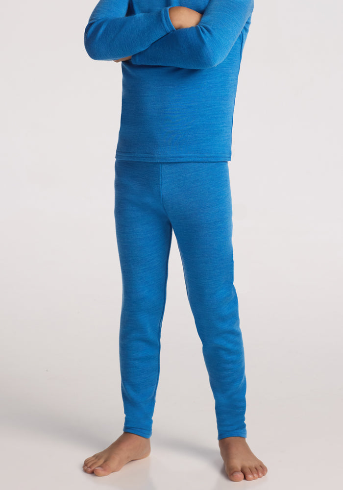 A child stands barefoot on a white background, arms crossed and head out of frame, wearing Woolx Kids Explorer Base Layer Bottoms in Blue Razz and a matching long-sleeved merino wool top. 