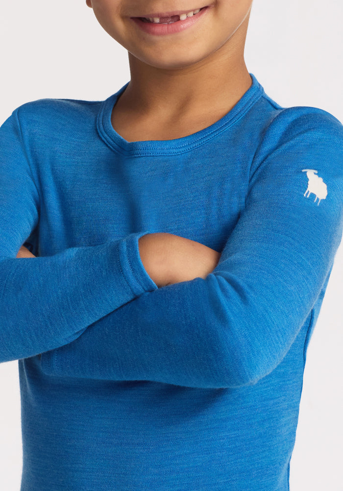 A child with arms crossed wears the Woolx Kids Explorer Base Layer Top in Blue Razz, featuring a small white sheep logo on the sleeve. Made from soft Australian Merino wool, only their lower face and torso are visible.