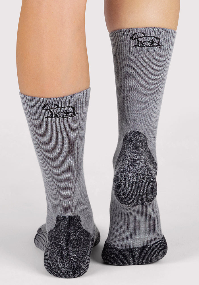 A person wears Woolx Womens Harbor Crew Sock - Grey, featuring moisture-wicking fabric, black textured heels and toes, and a black logo with text on the back near the top, standing on a light surface.