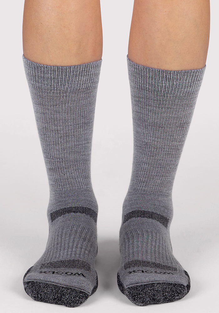 A pair of feet wear the Woolx Womens Harbor Crew Sock in grey, featuring ribbed Merino wool with darker gray accents on toes, heels, and arch, standing on a light surface.