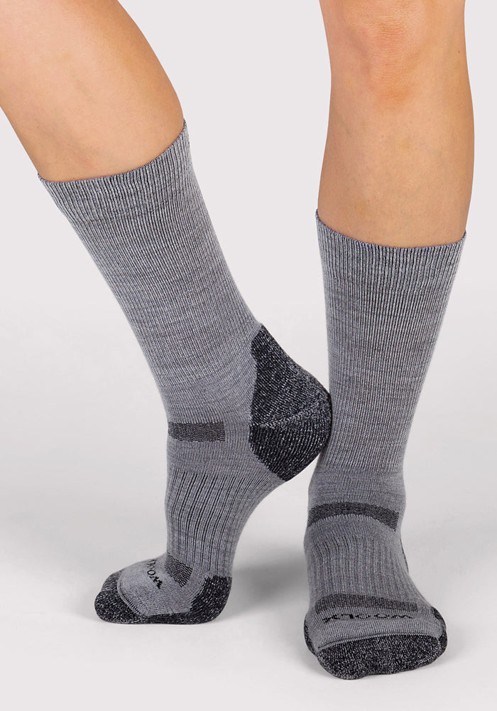 A person stands on a light surface wearing Woolx Womens Harbor Crew Socks in grey, featuring darker gray accents on the toes, heels, and arch, with one foot slightly raised and toes pointed downward.