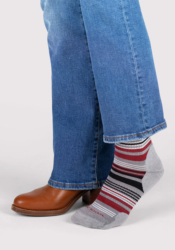 A woman steps out in blue jeans and brown shoes, revealing Woolx Womens Everyday Crew Socks in Light Grey Stripe—crafted from moisture-wicking, odor-resistant Merino wool with red, black, and white accents.