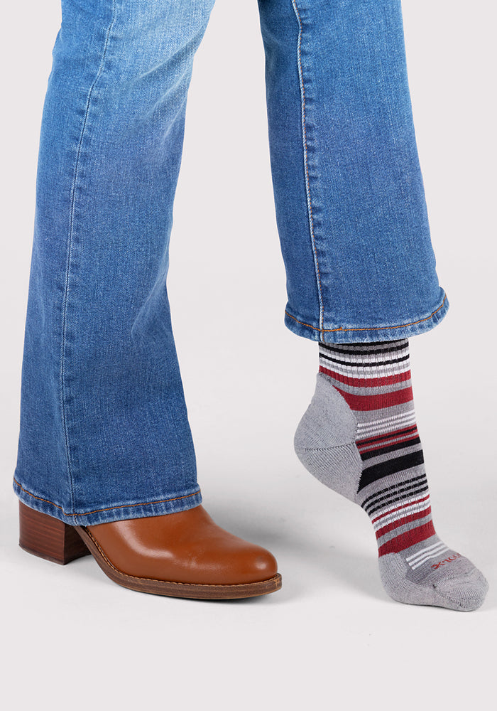 A person in blue jeans stands with one foot in a brown heeled shoe and the other in a Woolx Womens Everyday Crew Sock in Light Grey Stripe. The Merino wool sock features red, black, and white stripes. The background is plain white.