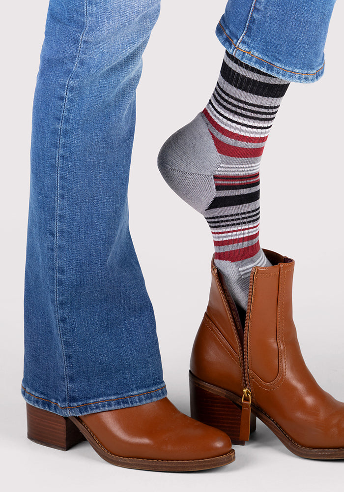 A woman in blue jeans slips off a brown ankle boot, revealing her foot in a colorful, odor-resistant Woolx Womens Everyday Crew Sock in Light Grey Stripe; her other foot stays in a matching boot.