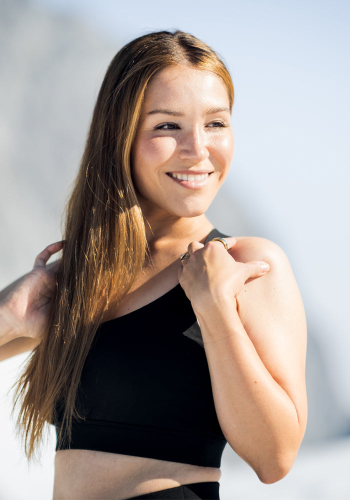 A woman with long, light brown hair smiles outdoors in athletic wear and the Woolx Livi Bra in black. Sunlight highlights her face as she holds her hair back, standing against a bright, blurred background.
