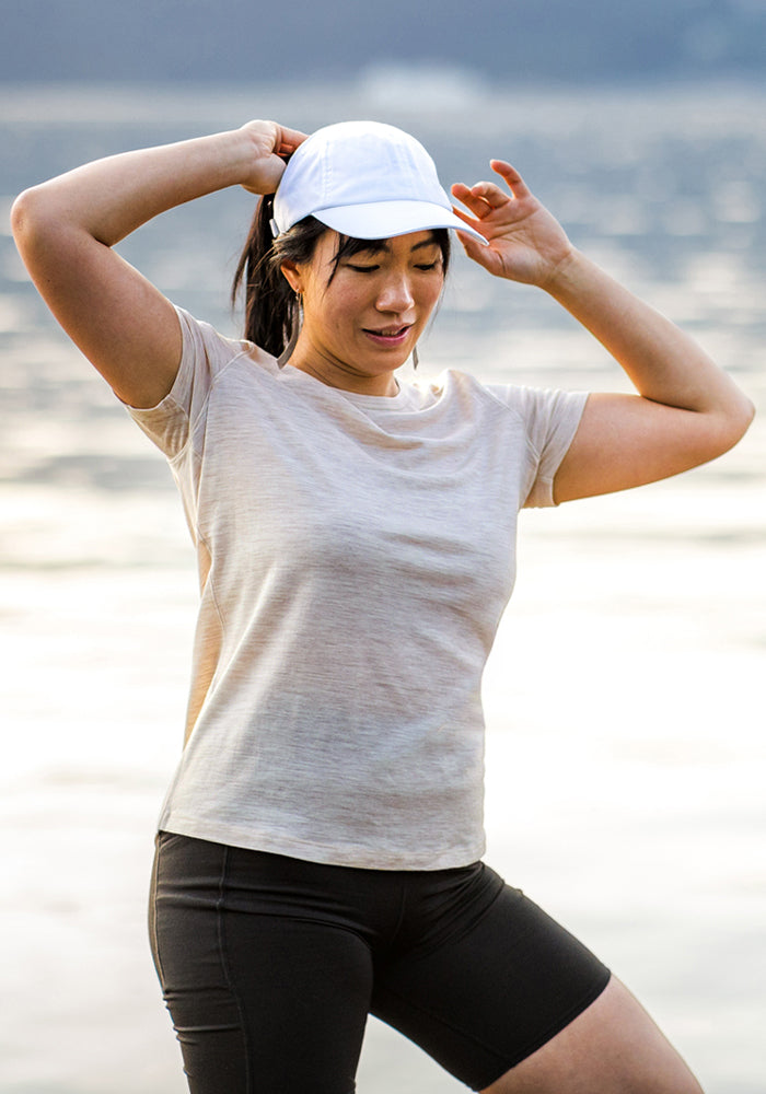 A woman in the Woolx Liza Crew Neck Tee - Cream Heather, a white cap, and black shorts stands by the water outdoors, adjusting her ponytail and smiling lightly.