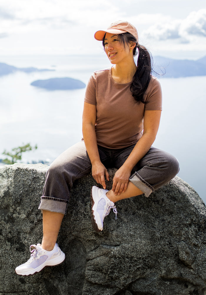 A woman in the Woolx Liza Crew Neck Tee - Sienna Kiss, rolled-up jeans, sneakers, and a peach cap sits smiling on a rock with mountains and water in the background under a bright sky.